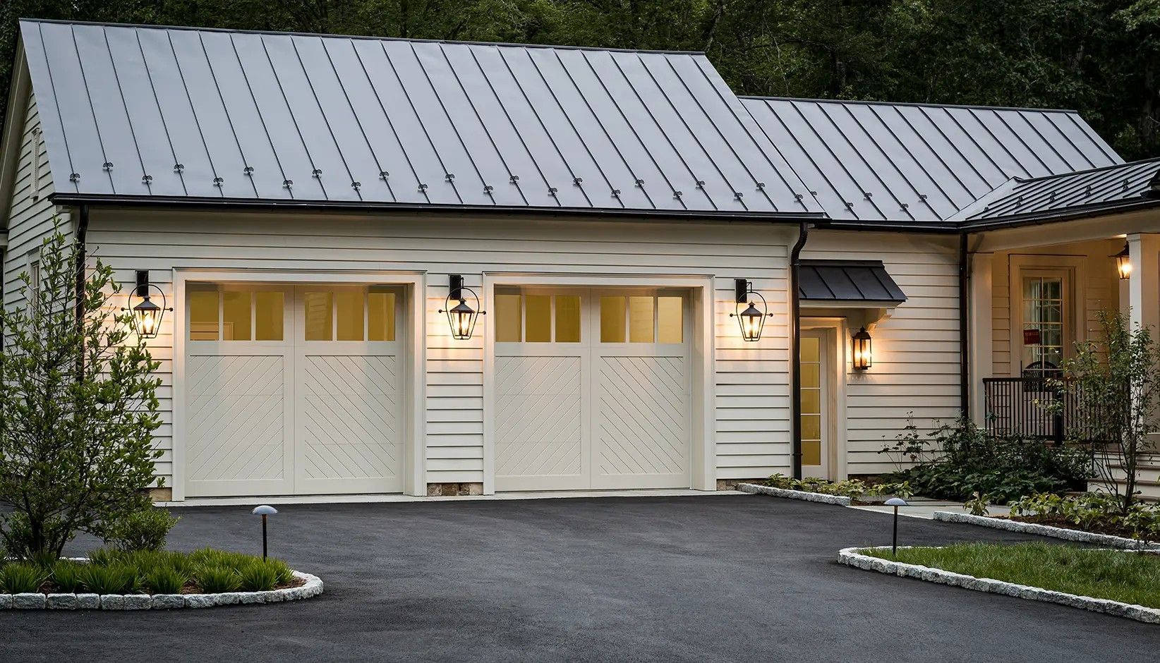 A white carriage-style garage with a metal roof and two garage doors, situated next to a house with a porch.