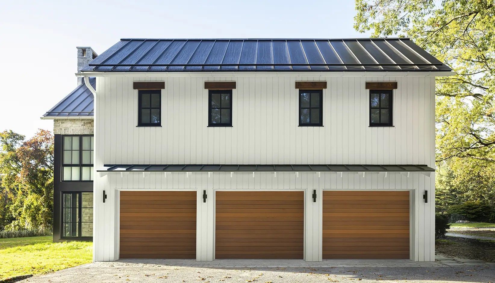 A white farmhouse-style structure with three wooden garage doors and a dark metal roof, set against a backdrop of trees.