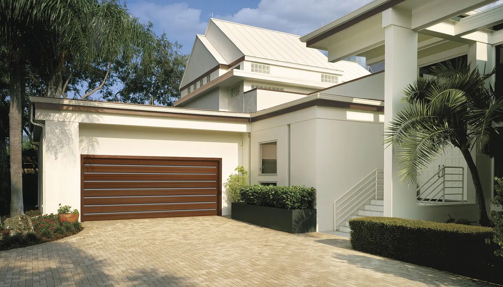 A modern, white multi-story home with a wide brown garage door, gravel driveway, and landscaped green shrubs.