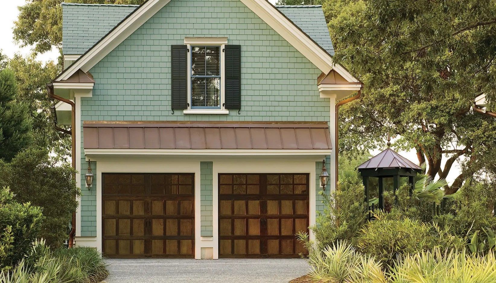 A light green two-story garage with two brown carriage-style doors, black shutters, and surrounding trees.
