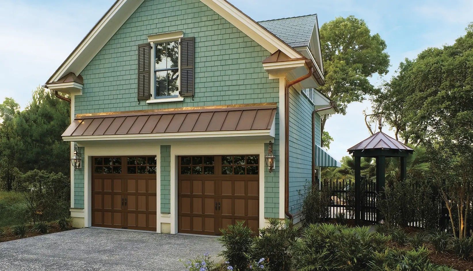 A two-story, green-sided garage with a copper roof and two wooden doors, set amidst lush trees and a stone driveway.