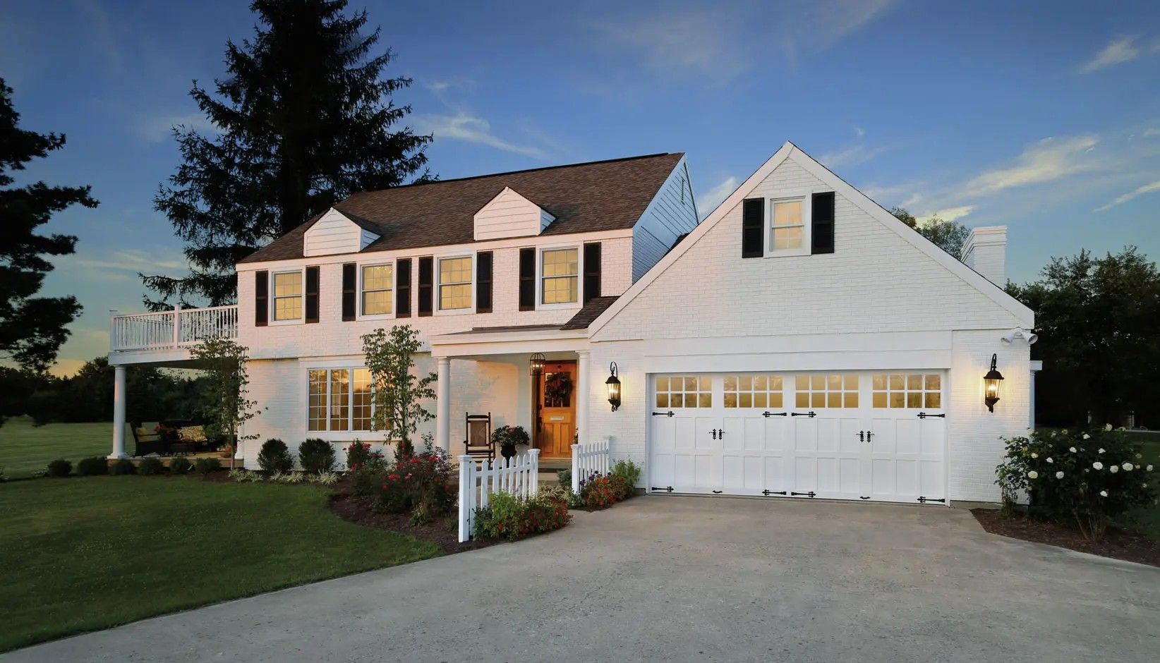 Two-story white house with black shutters, a two-car garage, a wooden deck, and a white picket fence under a blue sky.