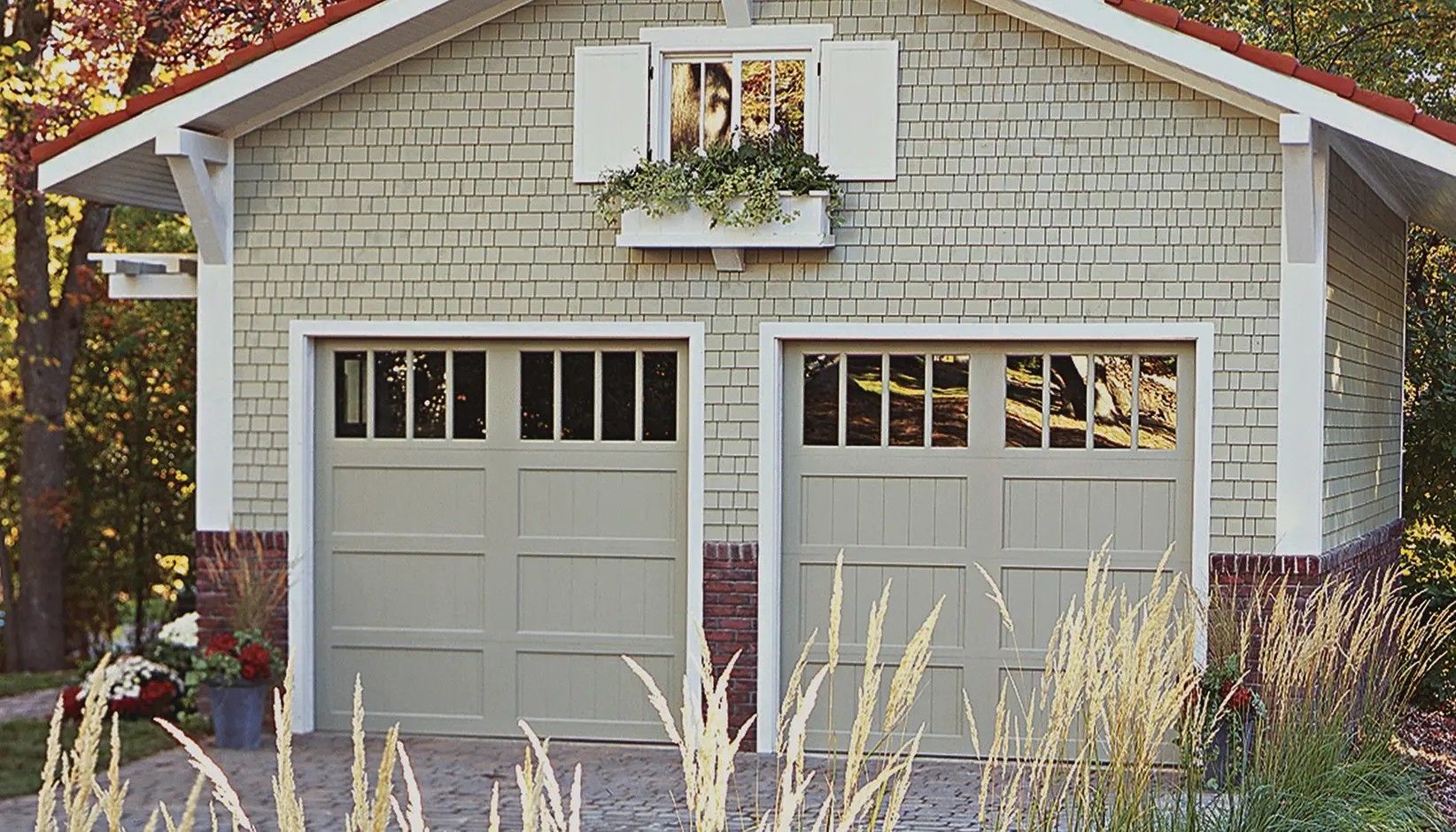 A detached, light-green two-car garage with white trim, a window box, and a brick base, set against a wooded backdrop.
