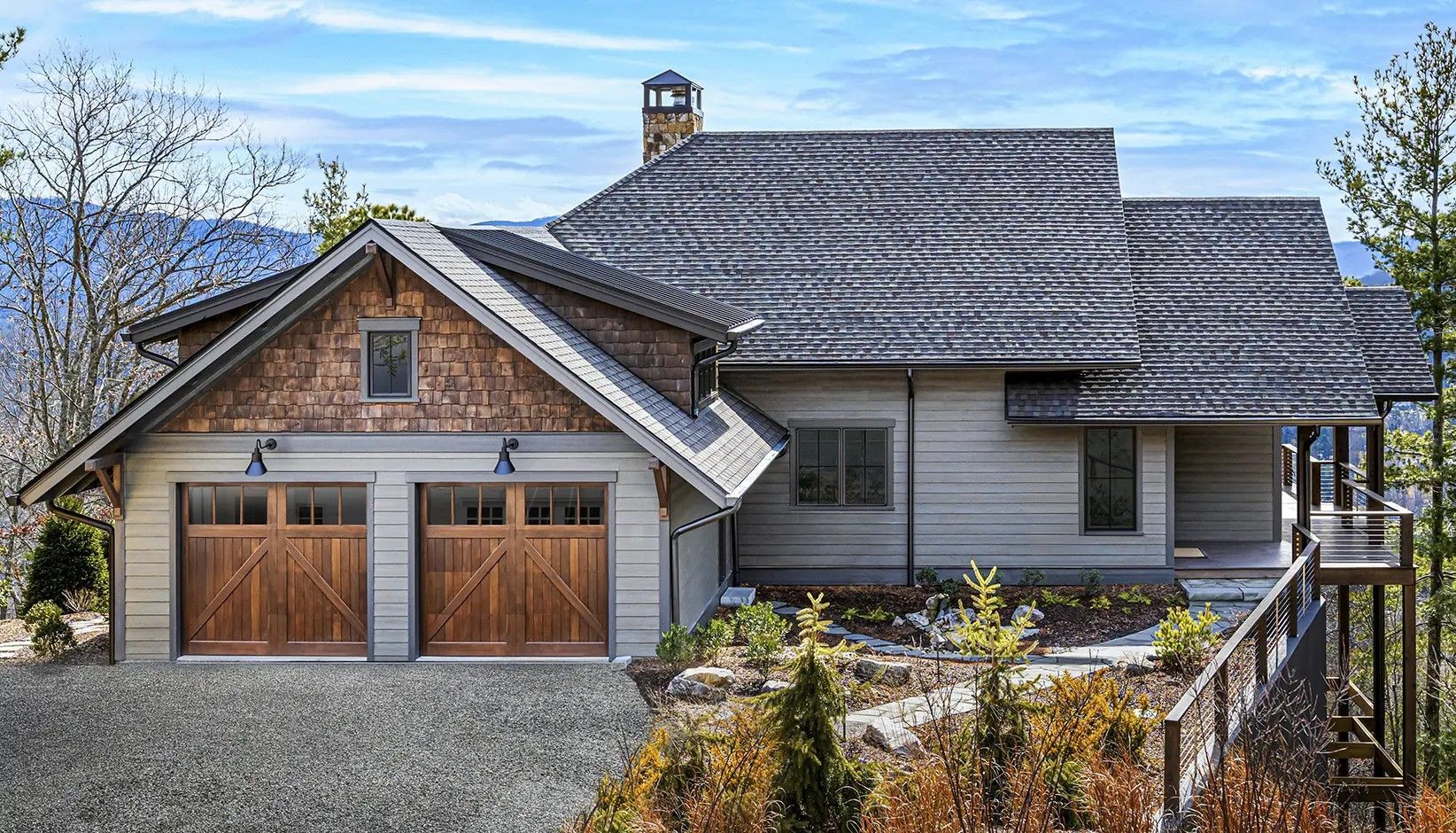 A two-story house with a wooden two-car garage, light siding, a shingled roof, and stone landscaping in a mountain setting.