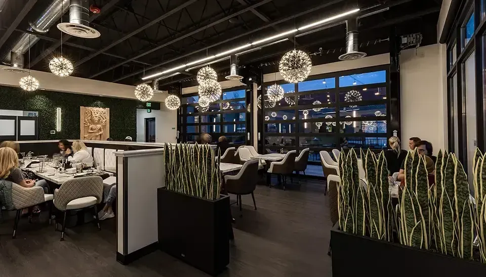 Modern restaurant dining room with white tables, hanging globe lights, and tall green plants by a windowed wall