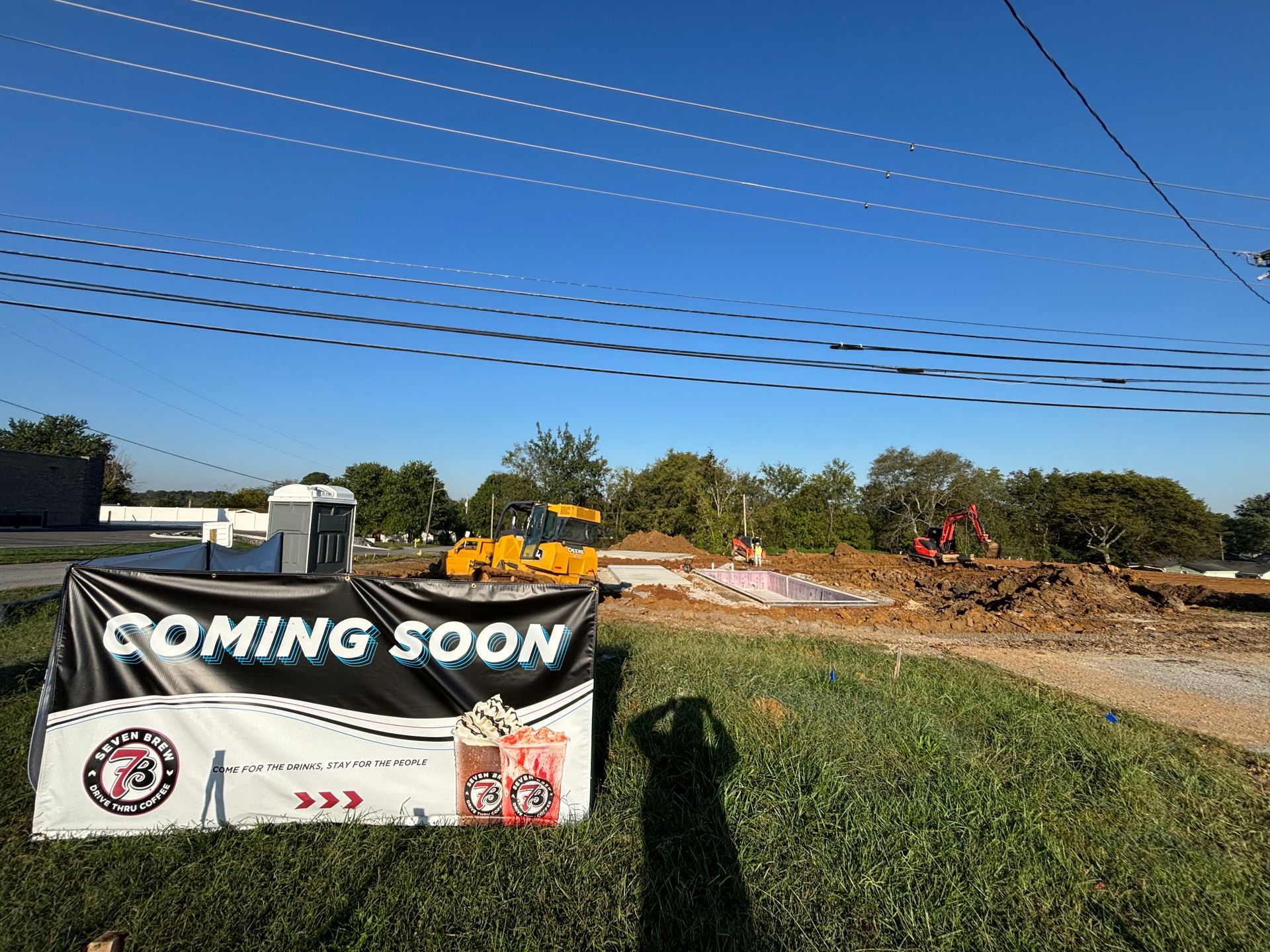 A coming soon sign is sitting in the grass in front of a construction site.