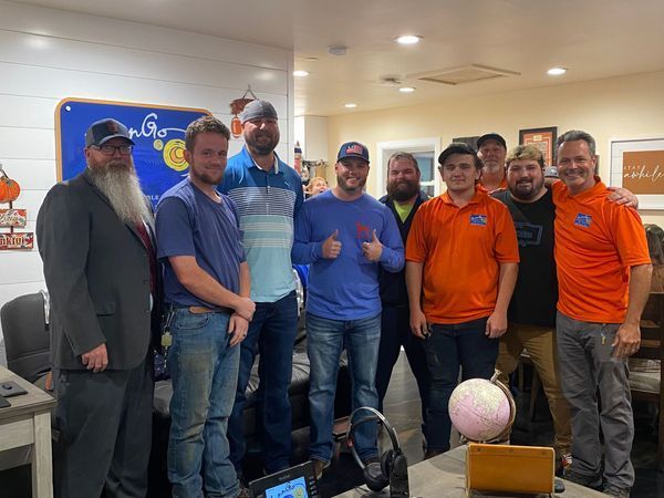 Group of men smiling indoors, gathered for a photo. Some wear orange or blue shirts. A globe is on a table.