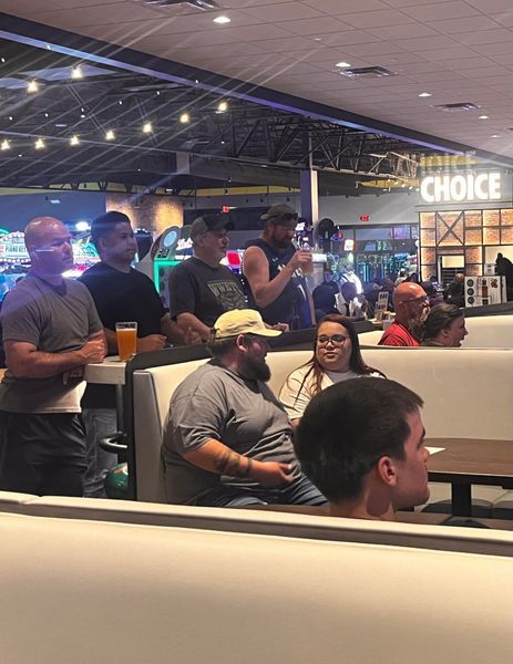 People sitting in a booth at a bowling alley, watching something. Some have drinks. Bright lighting.