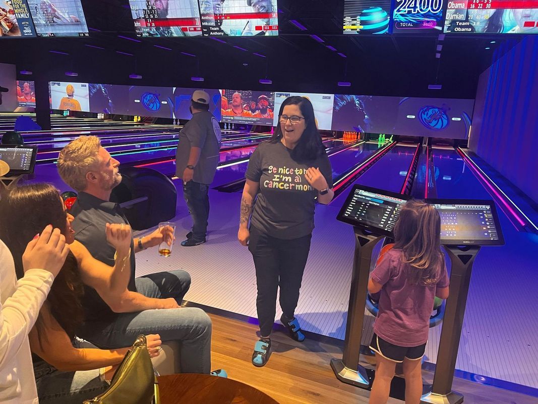 People bowling at an alley. A woman cheers, a child uses the control screen, others watch. Blue lanes, purple lights.