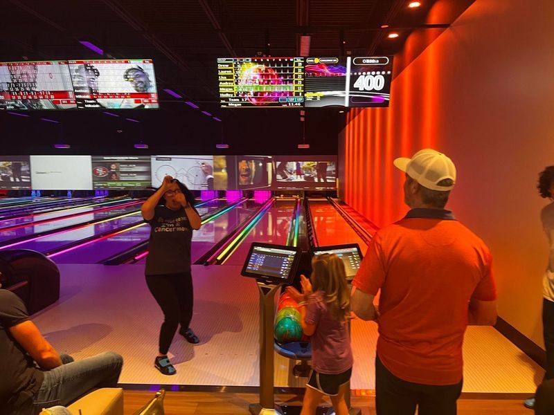 People bowling at a modern bowling alley. Woman celebrates, others watch, with colorful lane lights and screens.