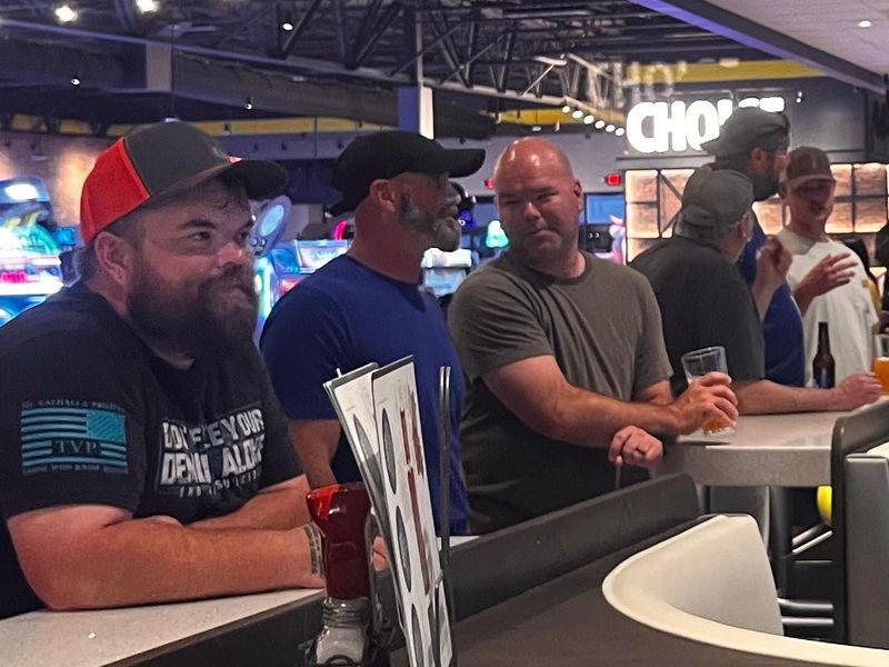 Men standing at a bar, some holding drinks, chatting. Casual setting with a 