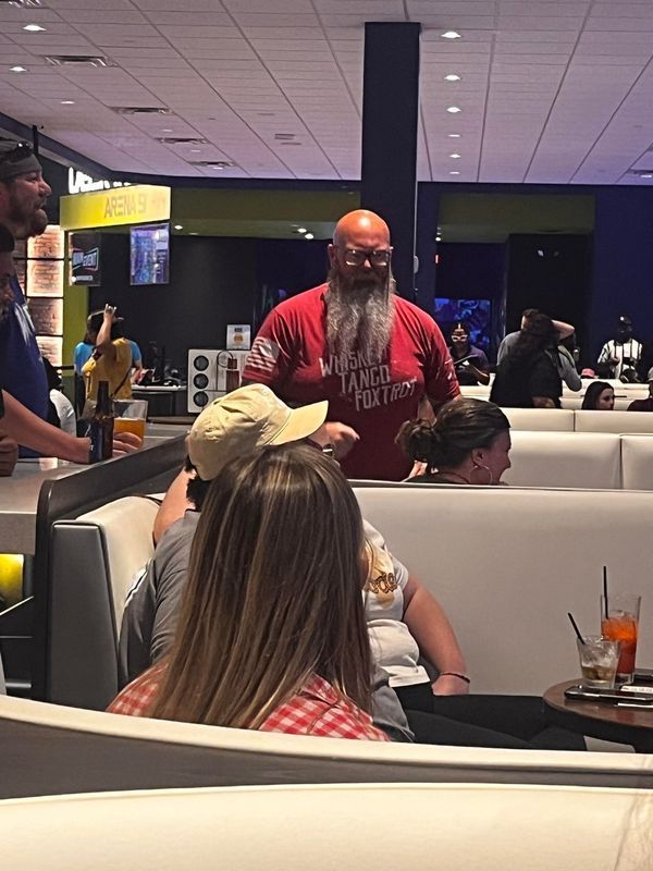 Man with long beard stands behind people seated in a bowling alley booth, wearing a red shirt.