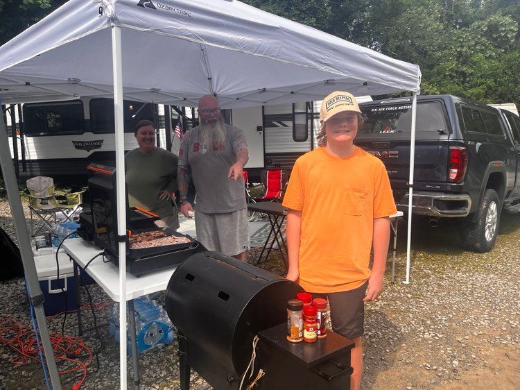 People grilling under a canopy at a campsite; a smoker, grill, and condiments are visible.