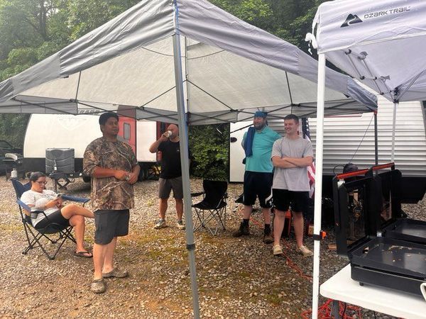 People under a canopy, camping, a grill, and a trailer in the background.