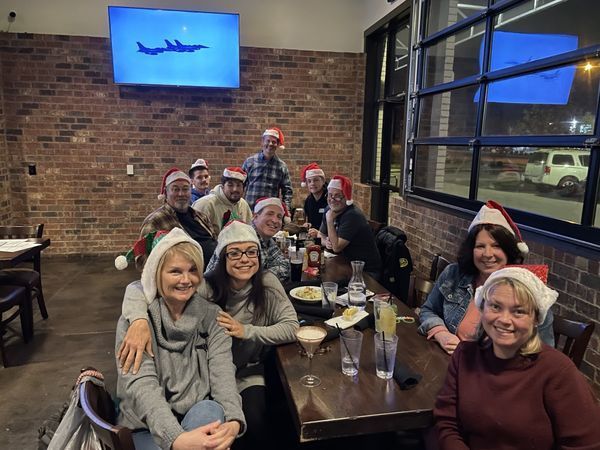 Group of people in Santa hats at a restaurant, gathered around tables, with brick walls and a TV screen.