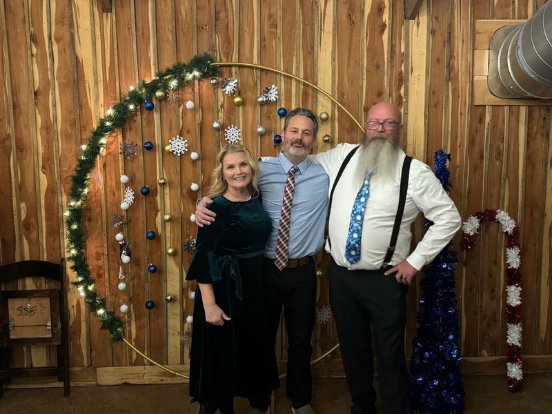 Three people pose in front of a circular holiday decoration. A woman in green, a man with a tie, and a bearded man smile.