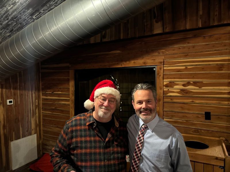 Two men smiling inside a rustic room; one wears a Santa hat, plaid shirt, the other a tie and button-down.