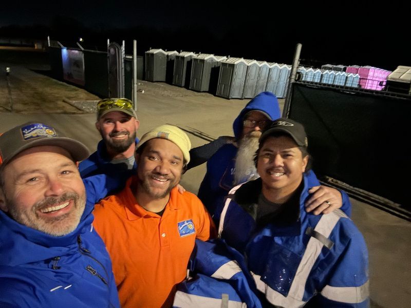 Group of five people smiling, in front of porta-potties at night, wearing work attire.