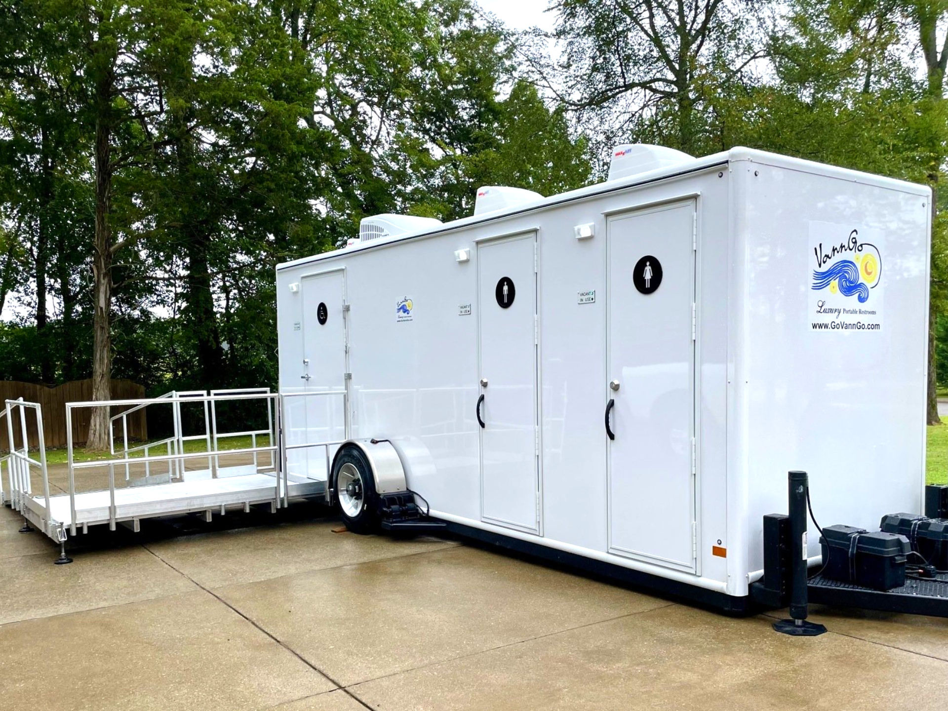 White trailer with portable restrooms, ramp, and logo, parked outdoors.