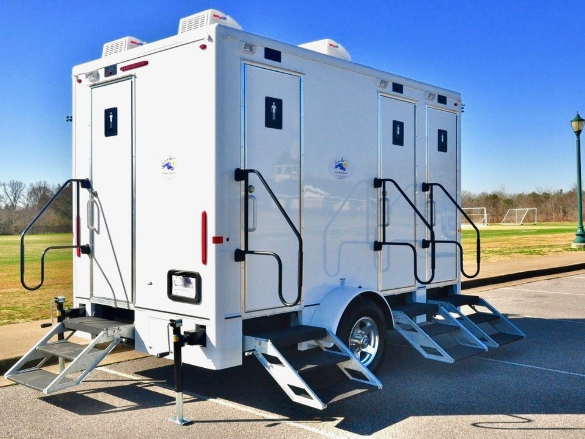 White portable restroom trailer with three stalls, ramps, and steps, parked outdoors.
