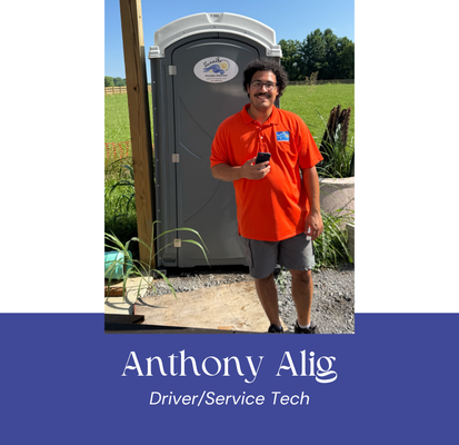 Man in orange shirt stands in front of a portable toilet. He holds a phone. Outdoors, sunny.