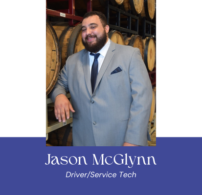 Jason McGlynn, Driver/Service Tech, poses by wooden barrels, wearing a gray suit and tie, smiling.