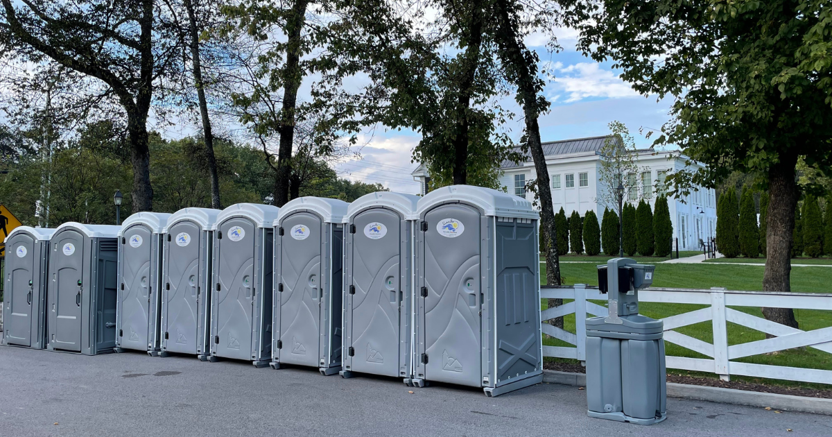 Row of gray portable toilets lined up on a street with a white fence, trees, and a building in the background.