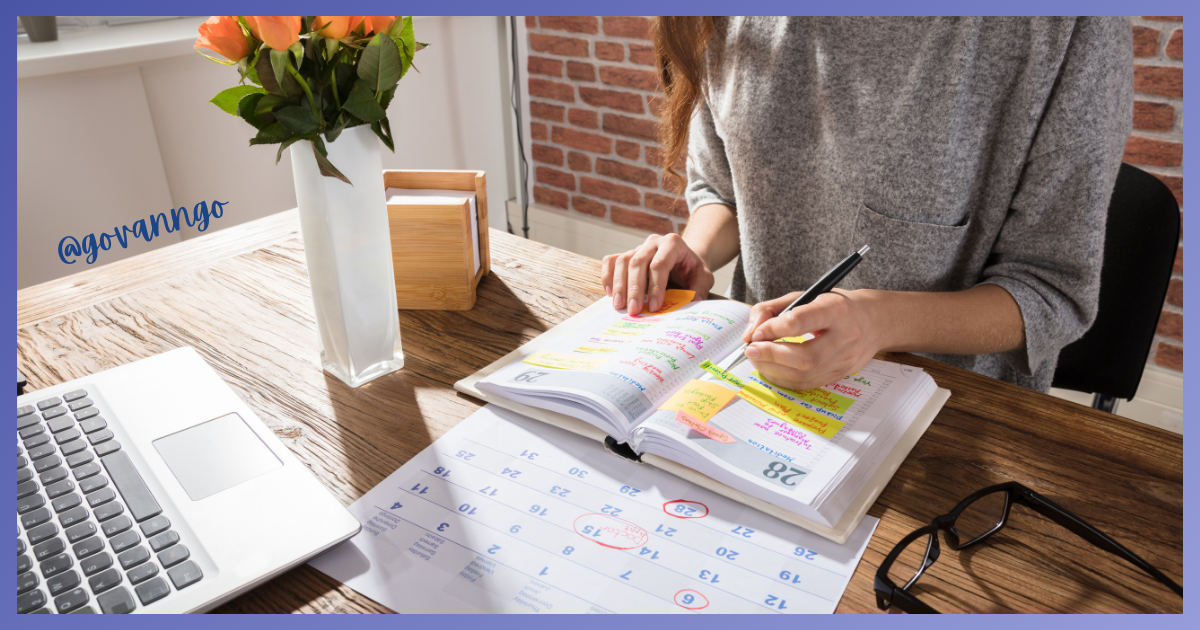 Woman writing in planner at desk with laptop, flowers, and calendar.
