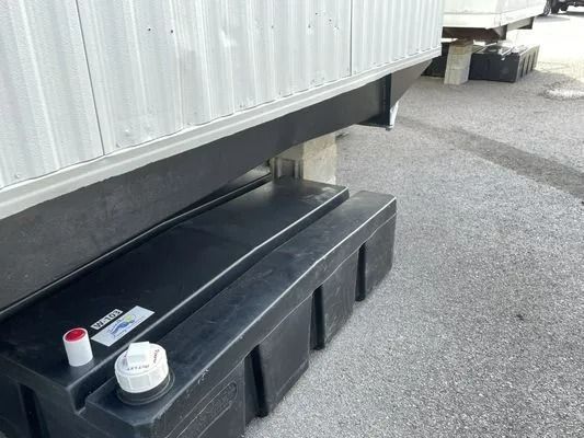 A long, rectangular black waste tank sits under the metal siding and frame of a mobile home on a gravel surface