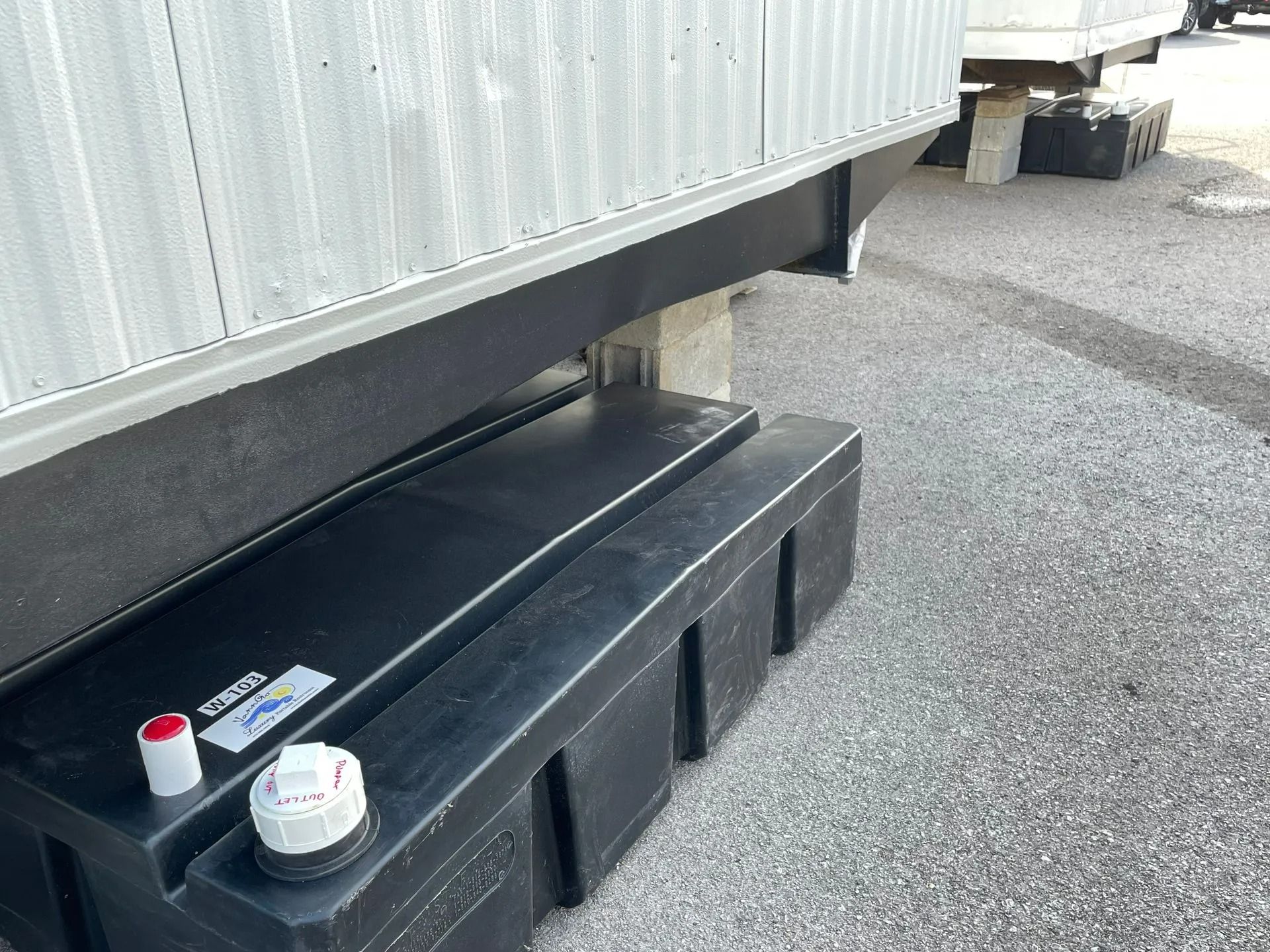 A black rectangular storage tank with two white caps sits on gravel beneath the edge of a mobile home structure