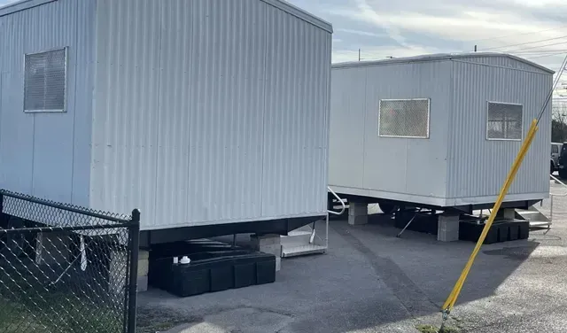 Two portable, light gray office trailers sit on raised foundations in a paved lot, viewed behind a chain-link fence
