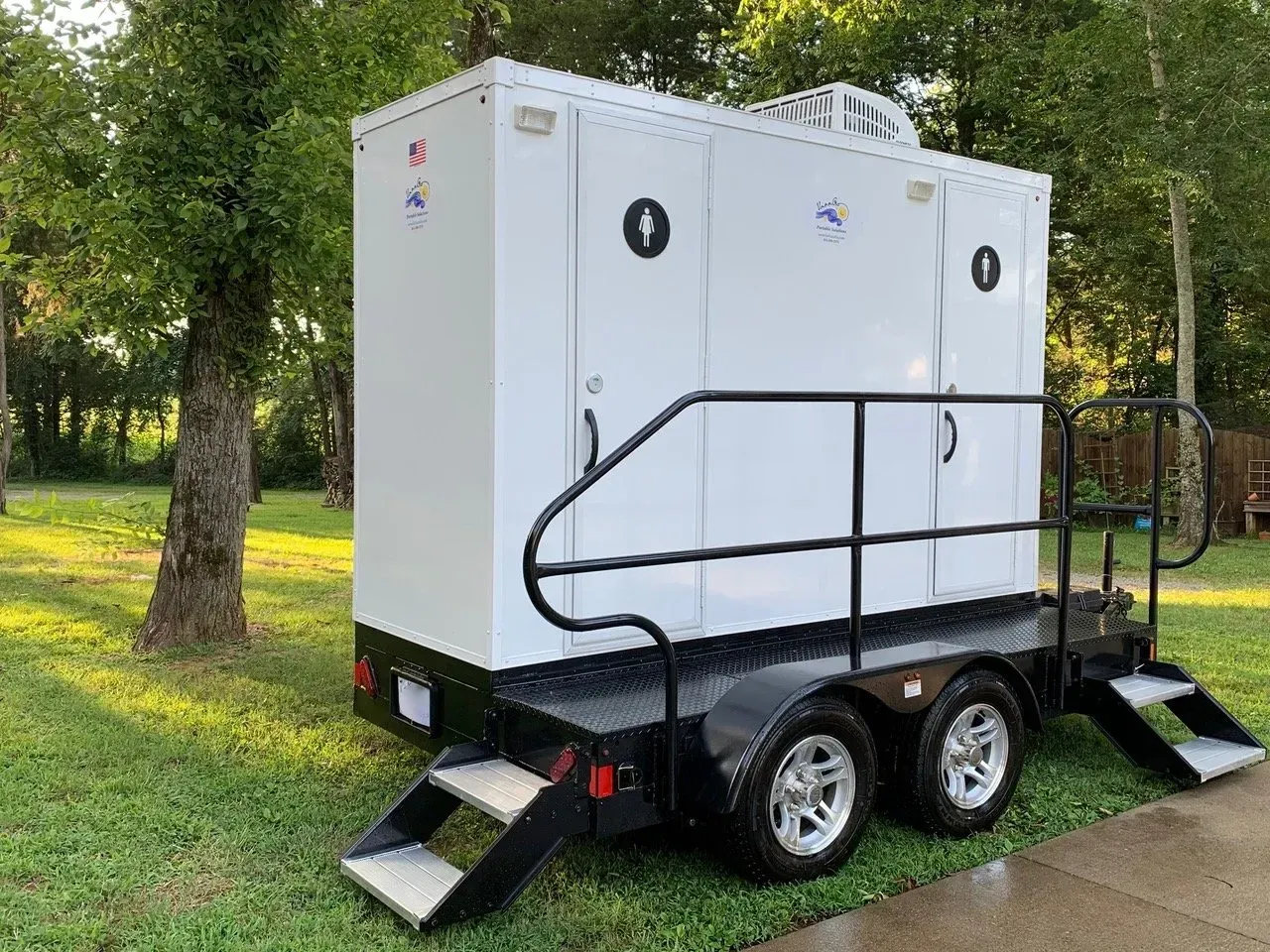 White portable restroom trailer on wheels with steps and railing, outdoors on grass.