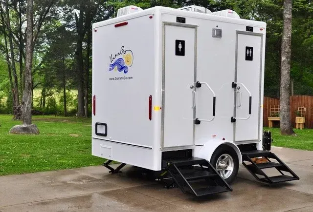 White portable restroom trailer with two separate stalls, one accessible, on a concrete pad with a grassy backdrop.
