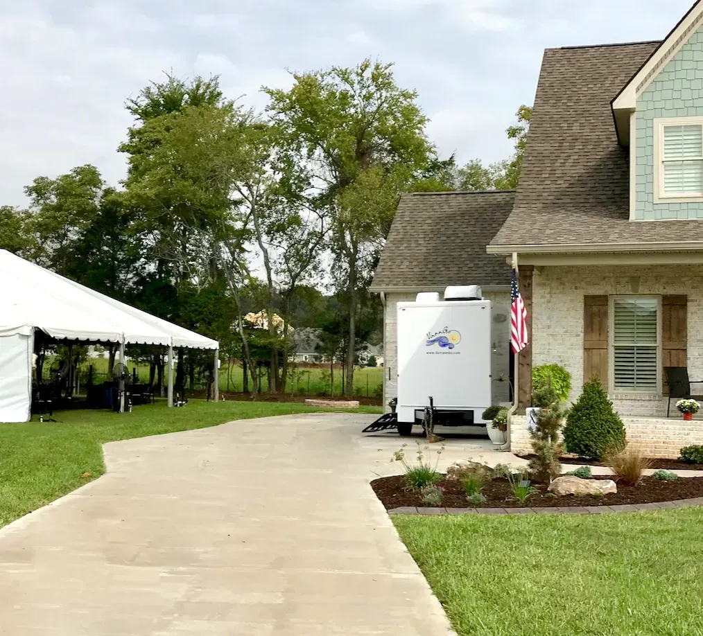 White tent and portable restroom trailer next to a house with a concrete driveway and lawn.