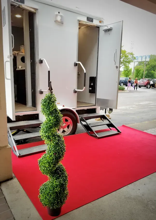 White luxury portable restroom trailer with open doors on red carpet, topiary, and ramp.