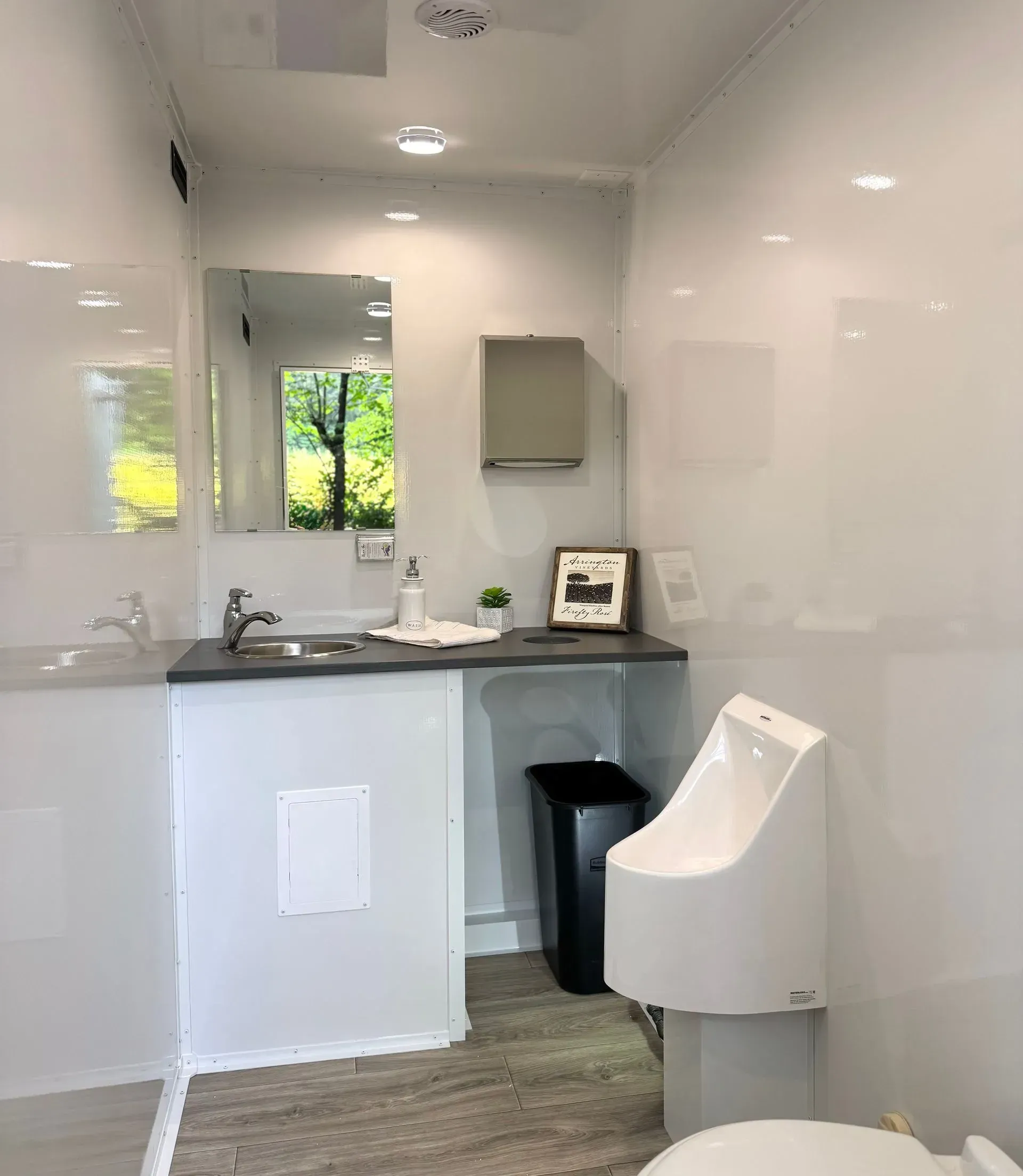 Modern white public restroom with sink, urinal, mirror, and trash can. Window view of trees.