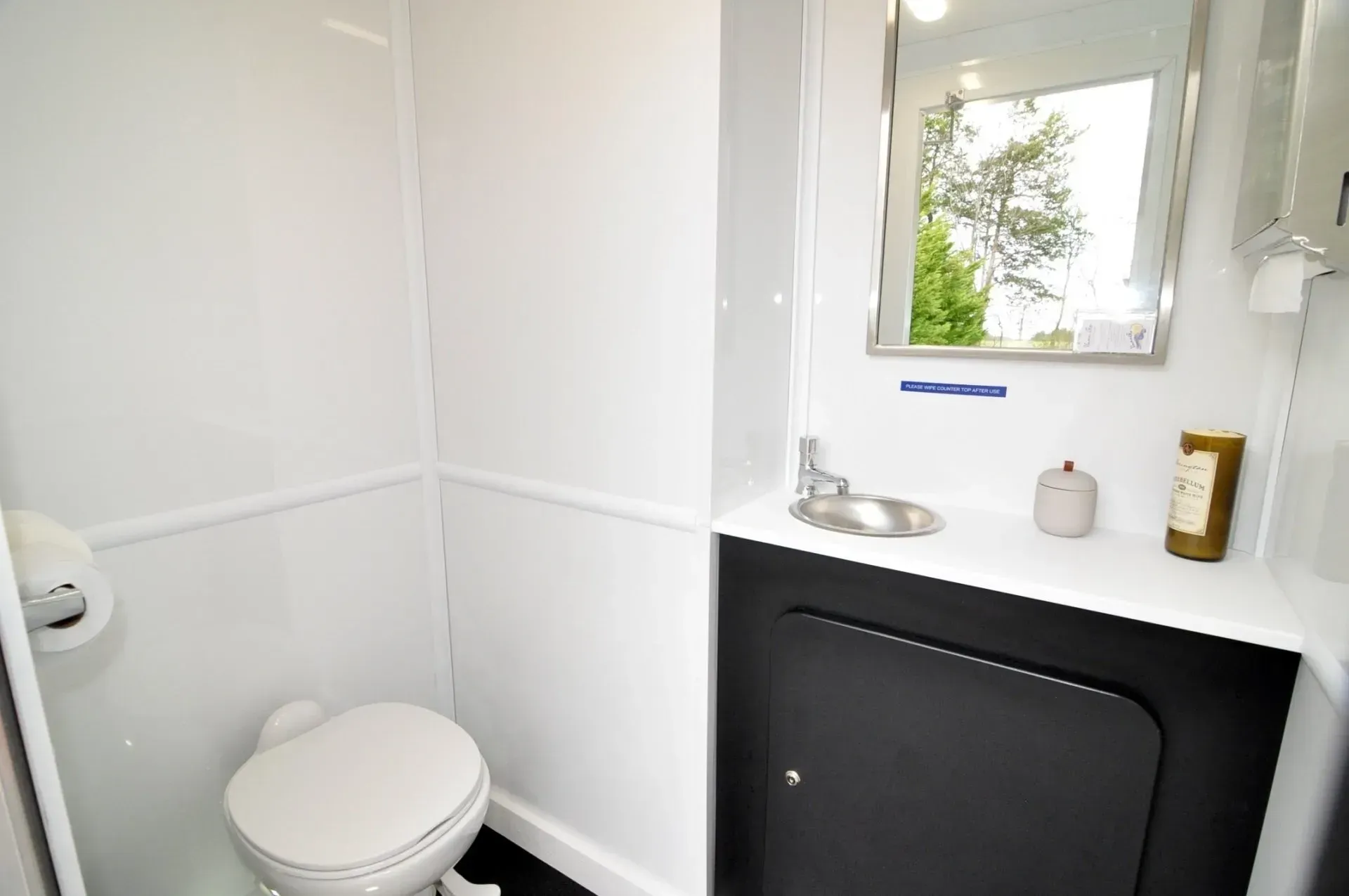 Interior of a portable toilet with a white wall, toilet, sink, and mirror.
