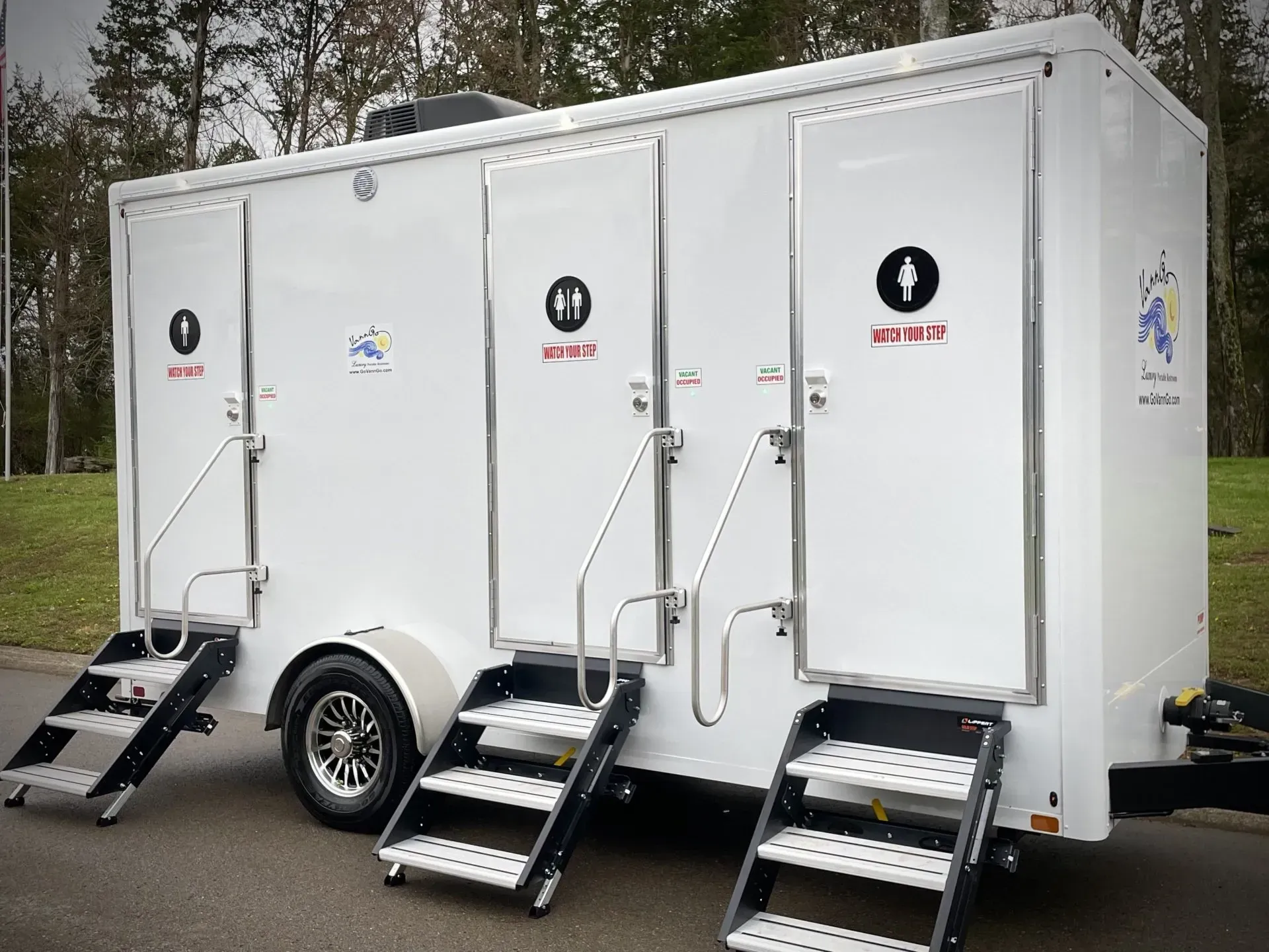 White portable restrooms with steps and gendered door signs on a trailer, parked outdoors.