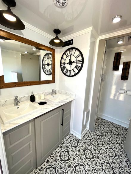 Bathroom interior with dual sinks, decorative clock, ornate patterned floor, and white walls.