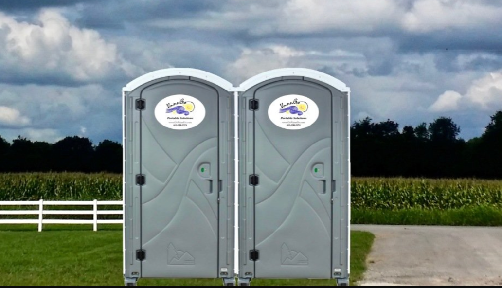 Two gray portable toilets on a rural roadside, against a cloudy sky and farmland.