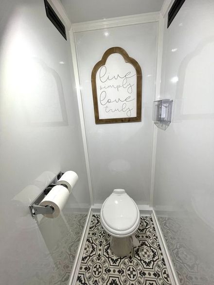 Small, modern restroom with white walls, patterned floor, and wooden sign that says, “Live simply, love deeply, care always.”