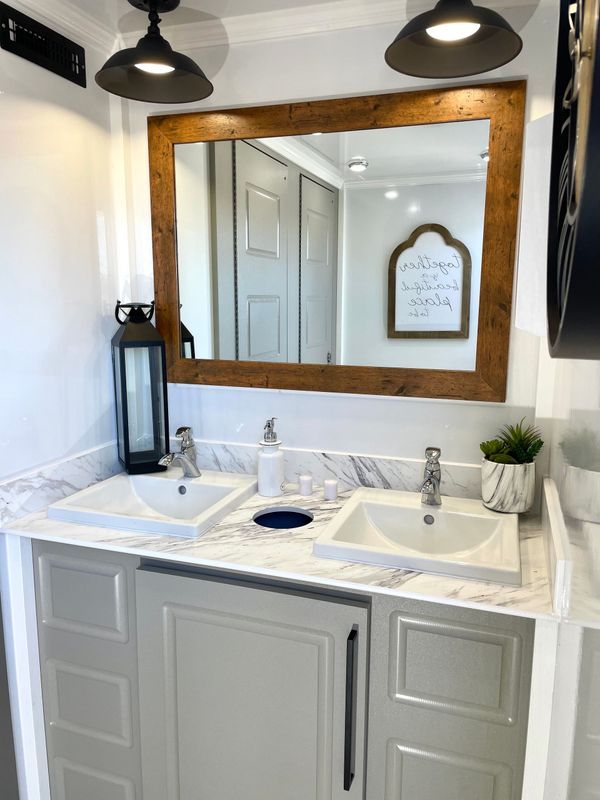 Bathroom interior with double sinks, marble countertop, wooden-framed mirror, and decorative lighting.