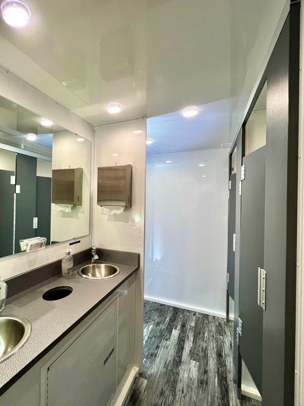 Interior of a modern portable restroom. Sinks, mirrors, and individual stalls with dark gray doors and gray/white walls.