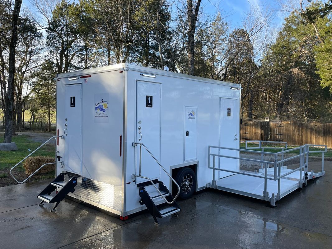 White portable restroom trailer with ramps, steps, and two restroom doors. Outdoors in front of trees.