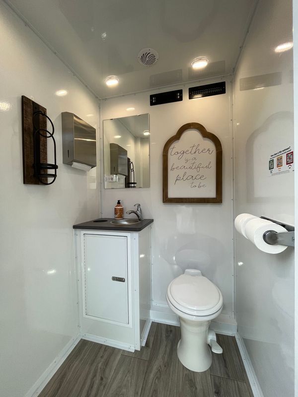 Interior view of a portable restroom with a toilet, sink, mirror, and decorative wall art in a clean, white space.