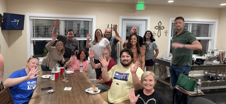 Group of people waving, gathered around a table indoors. Some have arms raised. The setting appears to be a gathering space.