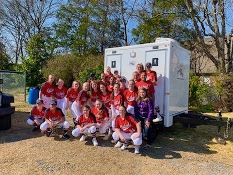 Softball team poses in red uniforms next to a portable restroom trailer.