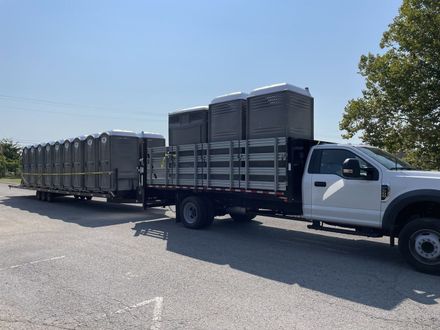 A truck towing a trailer, both loaded with portable toilets, parked on asphalt on a sunny day.
