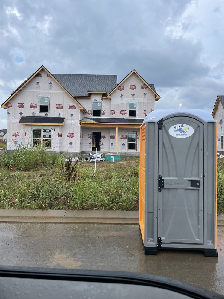 Gray portable toilet in front of a house under construction on a cloudy day.