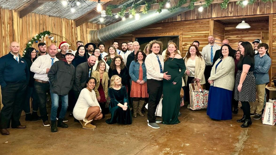 Group of people gathered inside a wooden-walled building. Several people are smiling. String lights and greenery overhead.